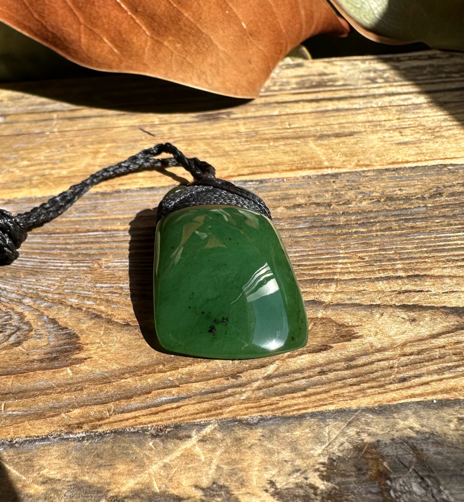 Green jade pendant with rounded rectangular shape on a black braided cord resting on a wooden surface
