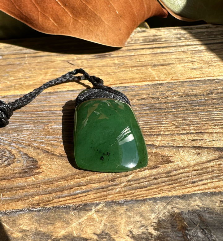 Green jade pendant with rounded rectangular shape on a black braided cord resting on a wooden surface