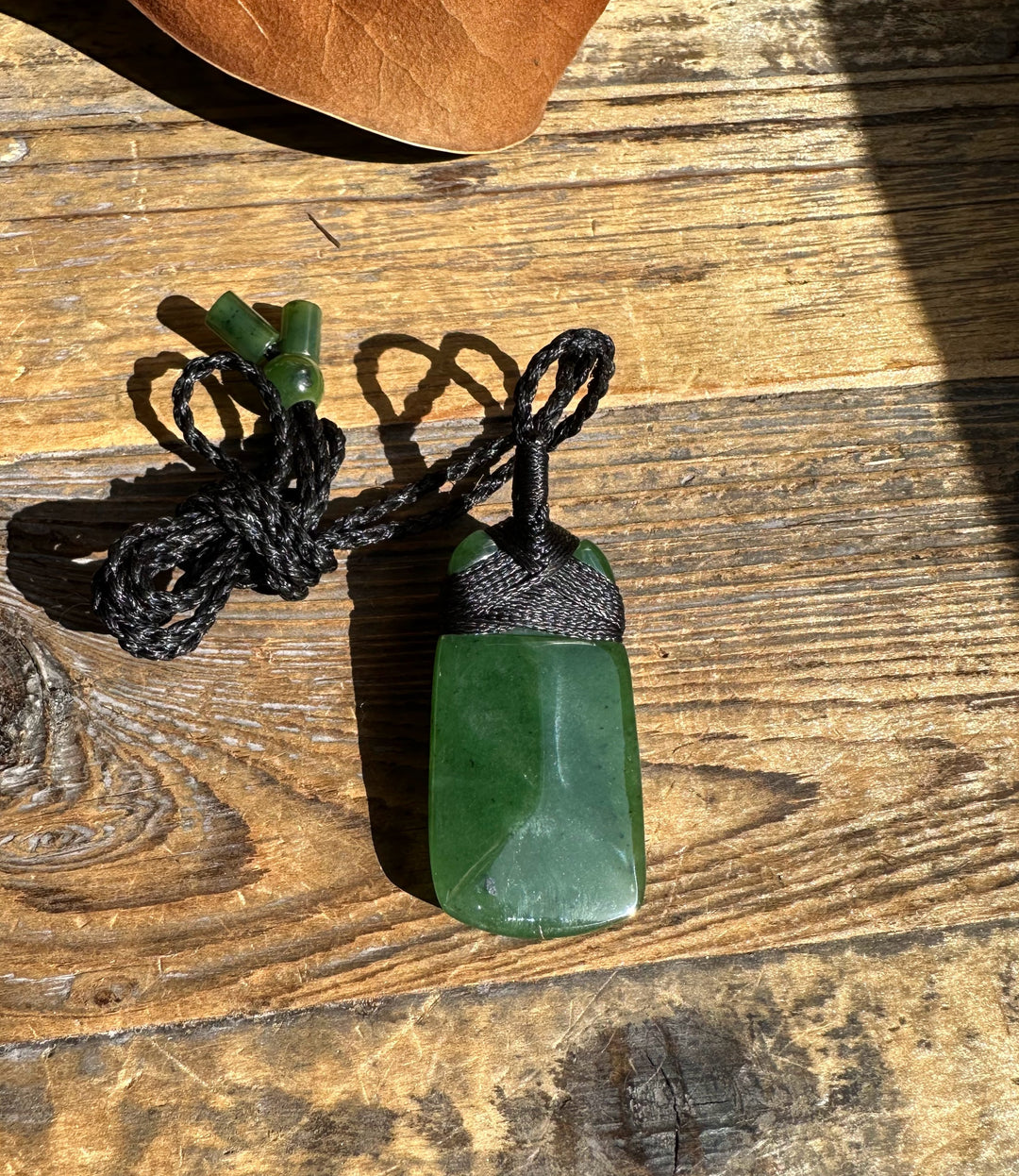 Green jade pendant with black woven cord and looped knot against sunlit wood