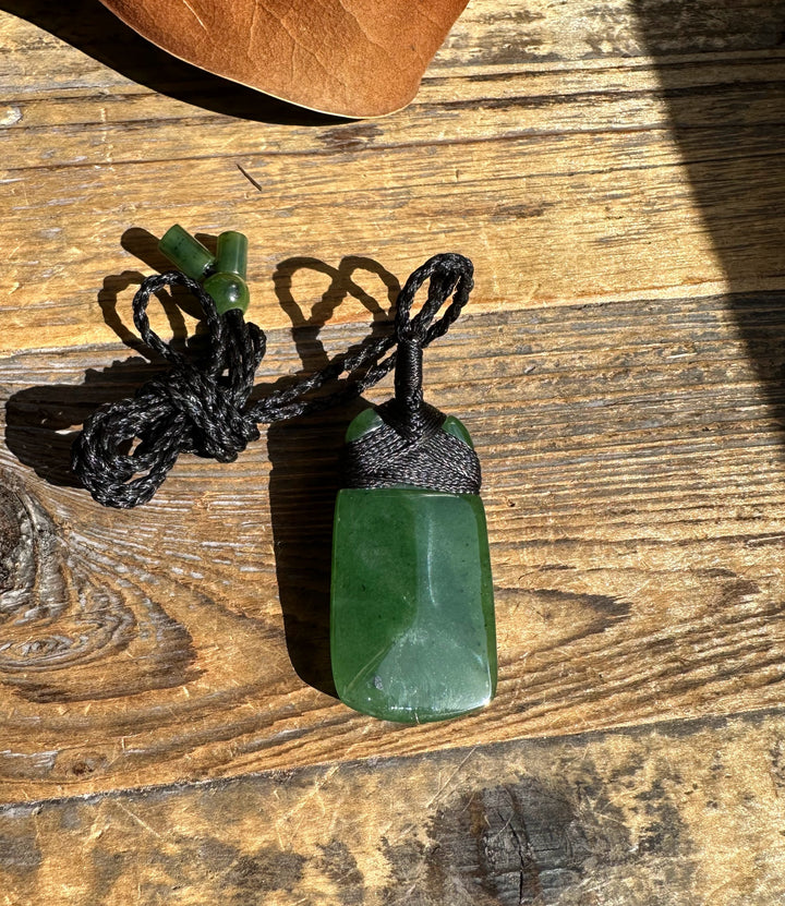 Green jade pendant with black woven cord and looped knot against sunlit wood