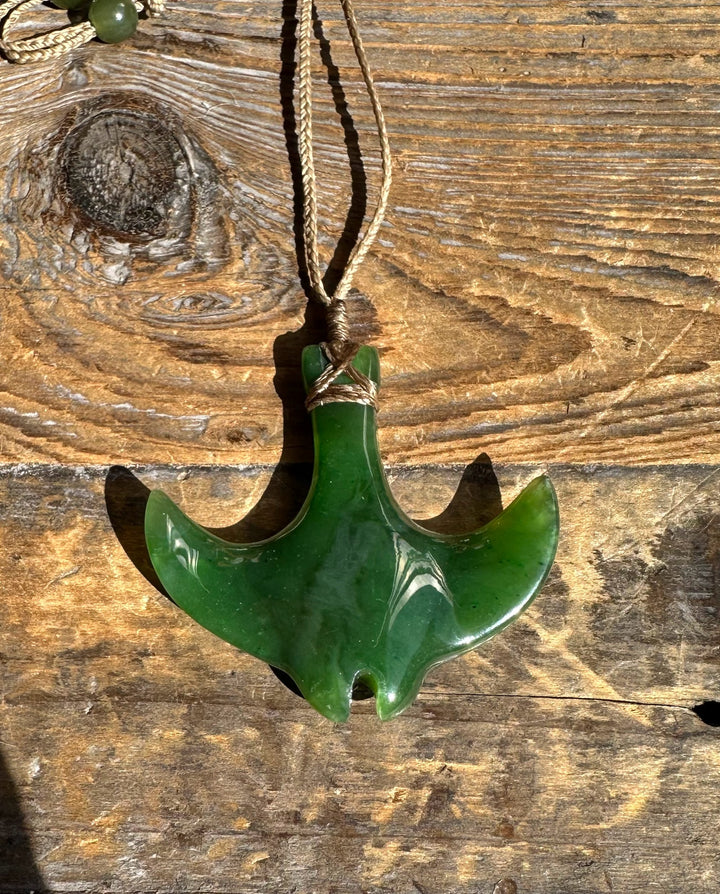 Green manta ray pendant carved from jade on beige braided cord necklace against wood background.