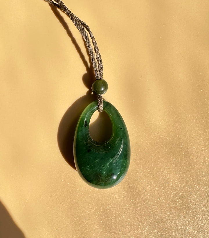Green jade hollow teardrop pendant on a braided cord with a bead against a tan background