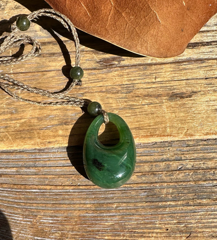 Donut shaped green jade pendant with a top hole on a braided cord with beads laid on wood