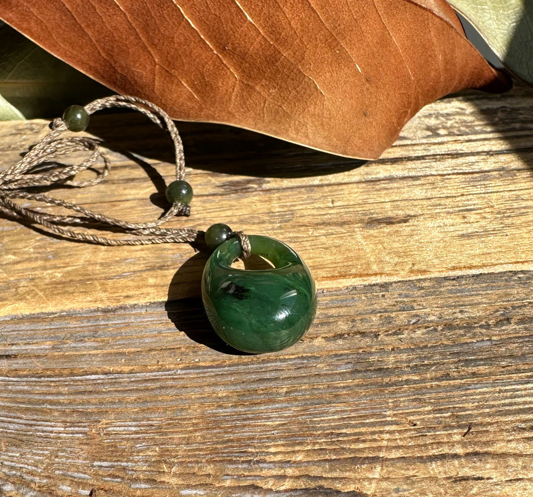 Jade donut pendant on a braided cord with green beads, laid on weathered wood with a brown leaf