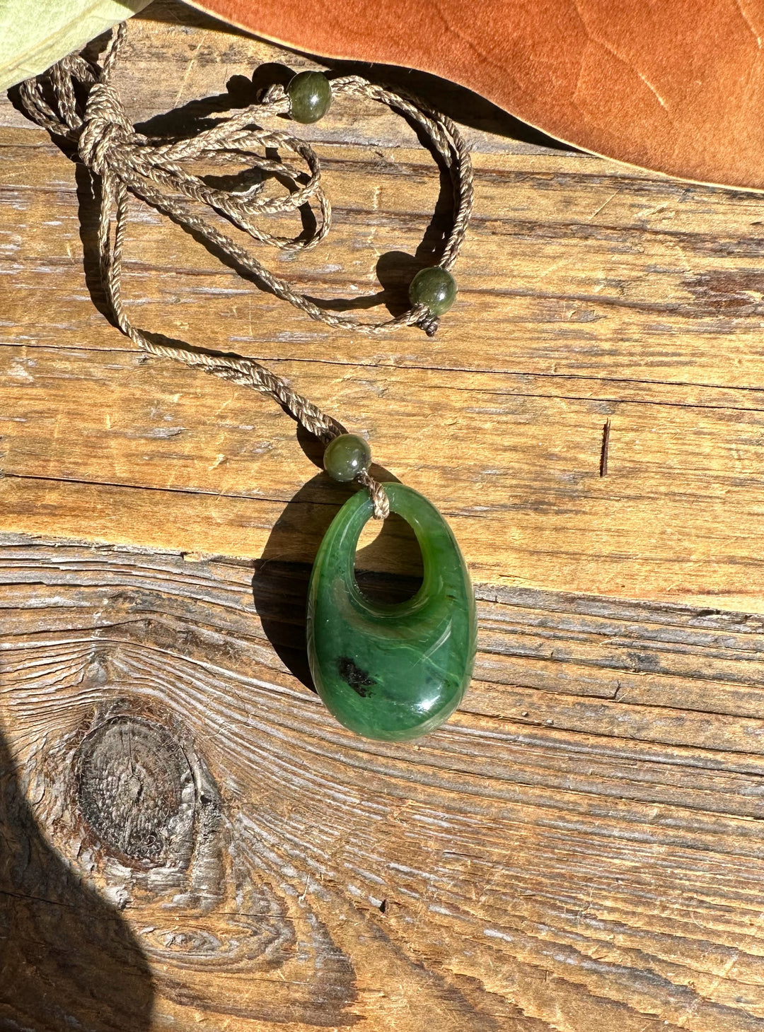 Green jade pendant with circular hole on a twisted cord resting on sunlit wooden surface.