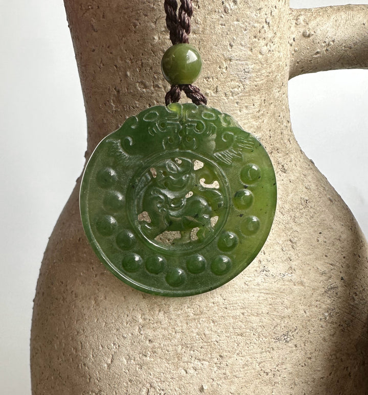Green jade Pixiu pendant circular disc with a central carved Pixiu and a dotted outer rim on a brown cord
