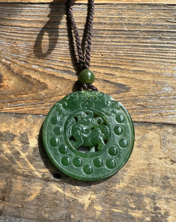 Front view of round green jade Pixiu pendant on a brown cord against a rustic wooden background