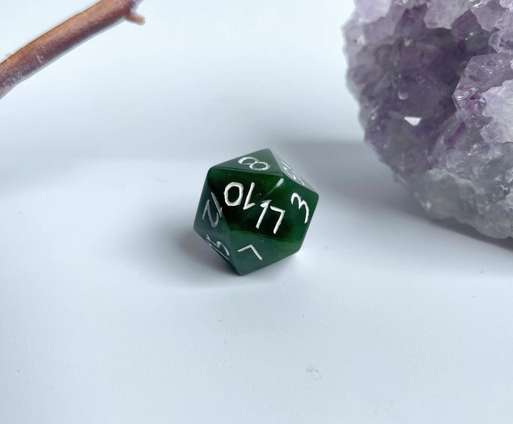 Green jade twenty sided die with white numbers on a pale surface beside a purple crystal cluster