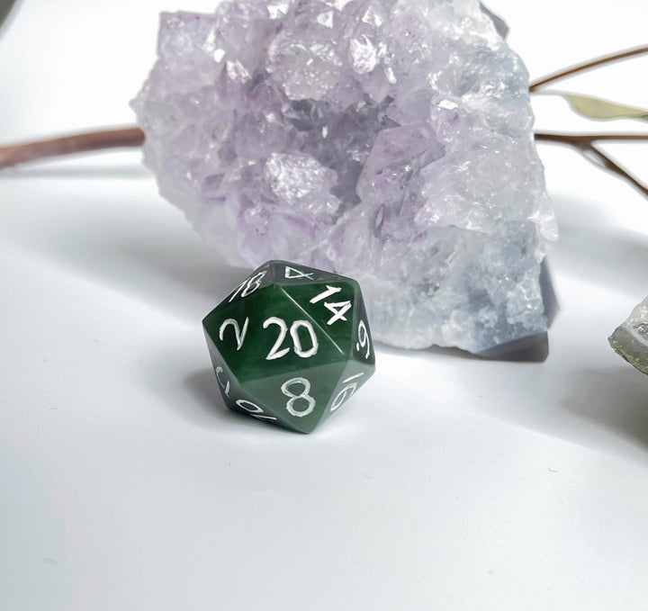 Jade green 20 sided die beside a purple amethyst cluster on a white surface.