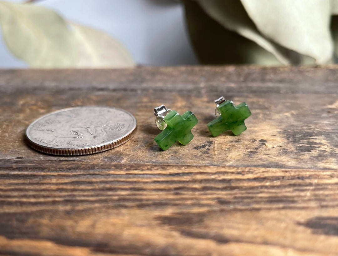 Jade cross earrings with silver posts placed beside a coin on a rustic wooden surface.