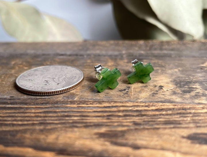 Jade cross earrings with silver posts placed beside a coin on a rustic wooden surface.