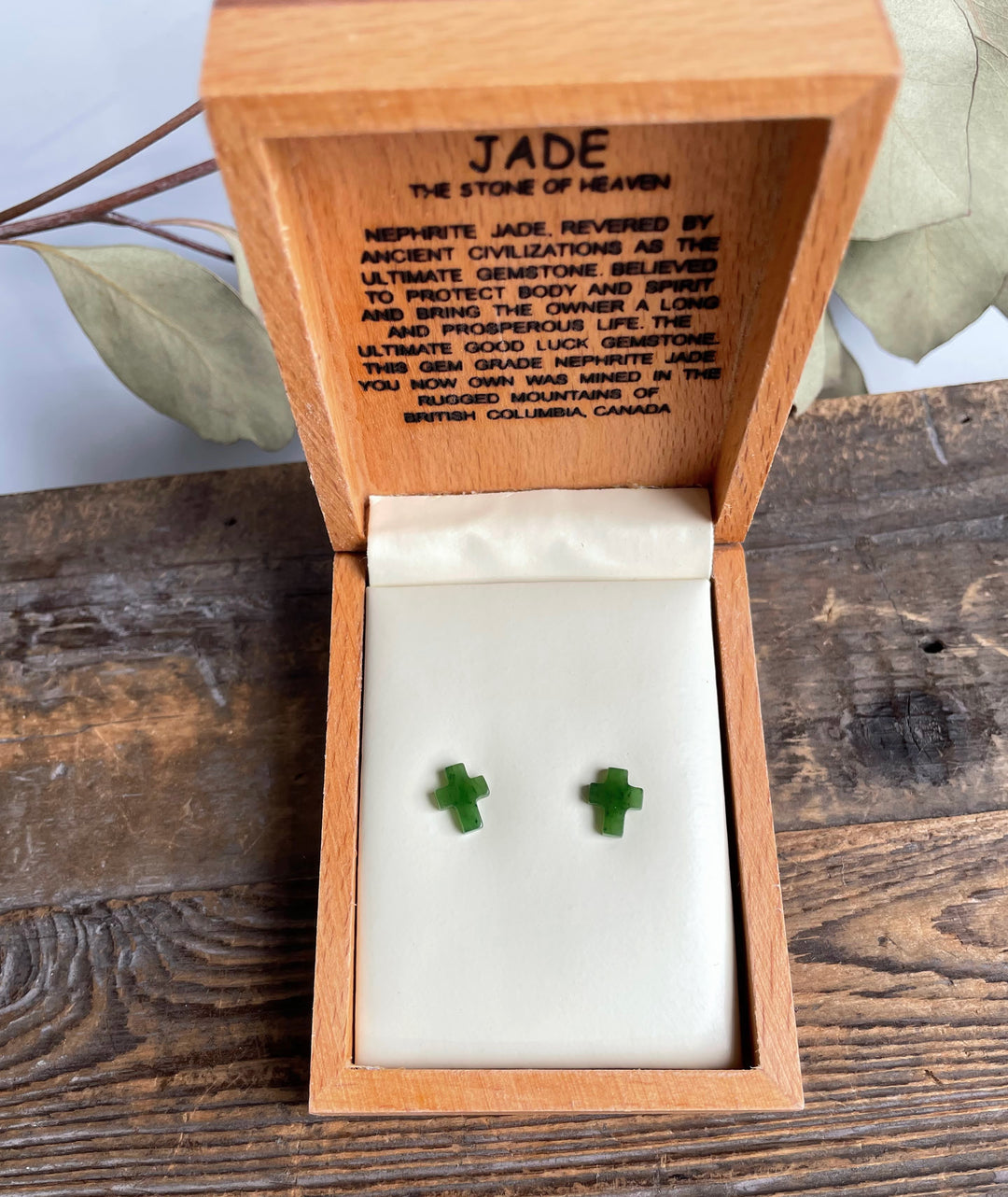 Open wooden box shows jade cross earrings on a white pad with an inscribed lid text.