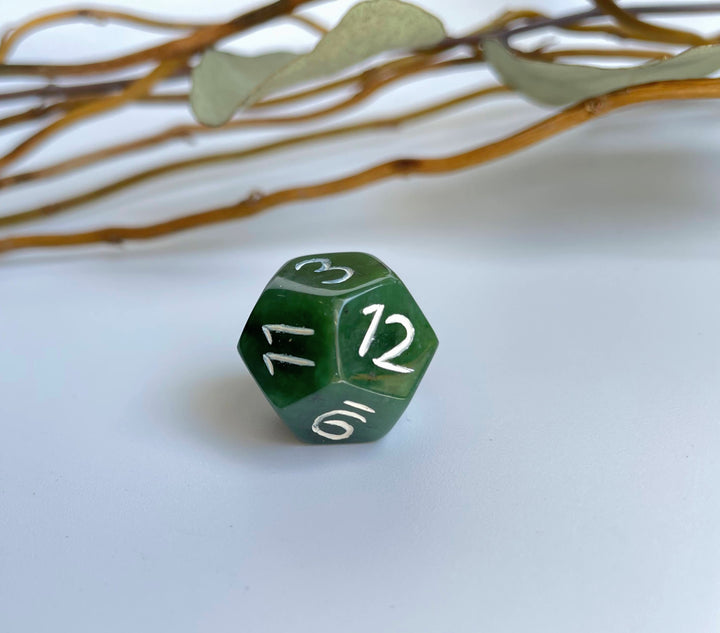 Green jade D12 die on a pale surface with decorative twigs in the background