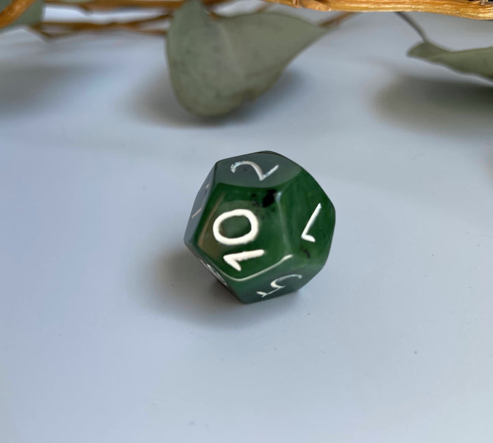 Green jade D12 die on a light surface with thin branches in the background