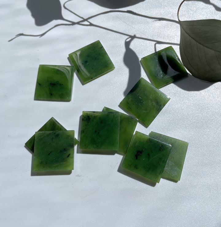 Green jade square cabochons on a white surface with shadows and a dried leaf