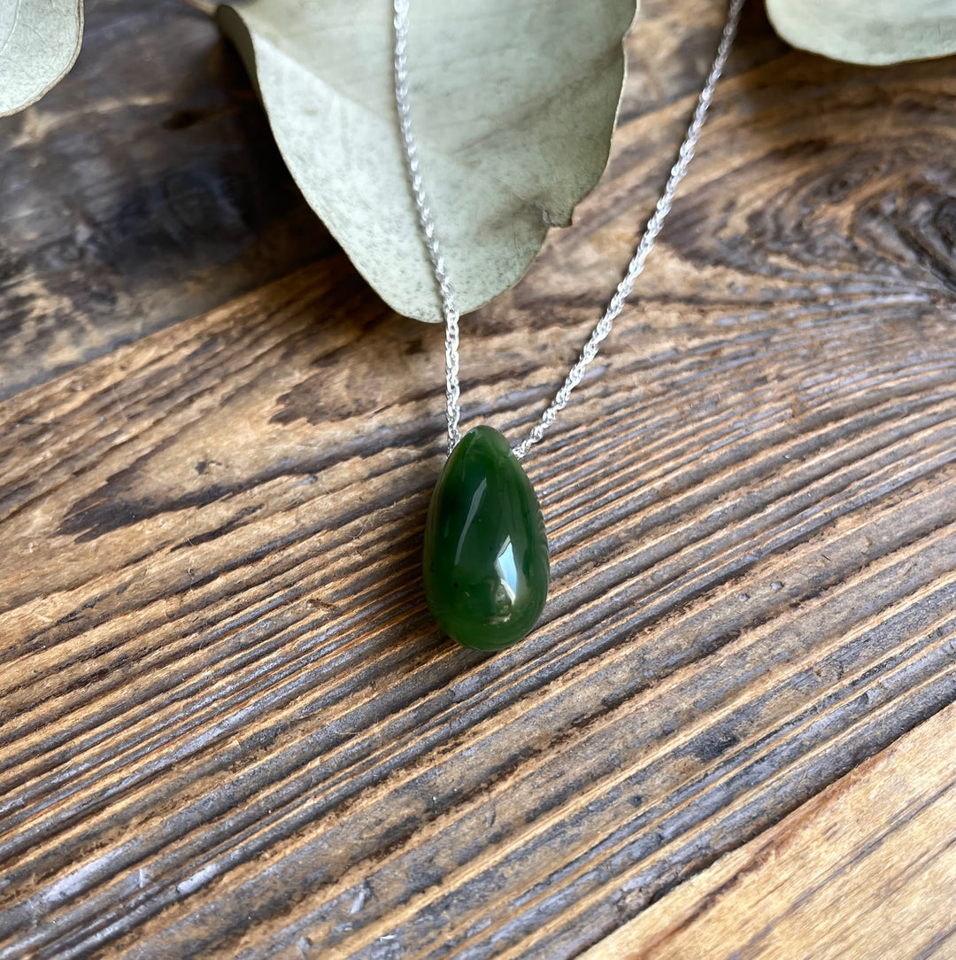 Close-up of a 20mm dark green Canadian Jade teardrop pendant on a silver chain against a wooden surface.