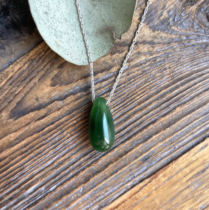 Dark green Canadian Jade teardrop pendant on a silver chain resting on rustic wood with eucalyptus leaves.