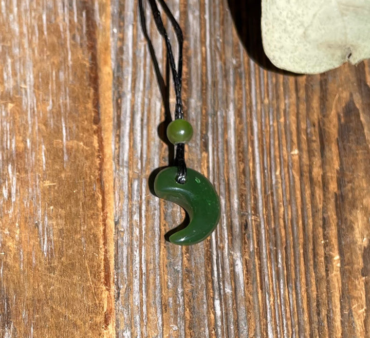 Dark green jade magatama crescent pendant with a small bead on a black cord against a wooden background.