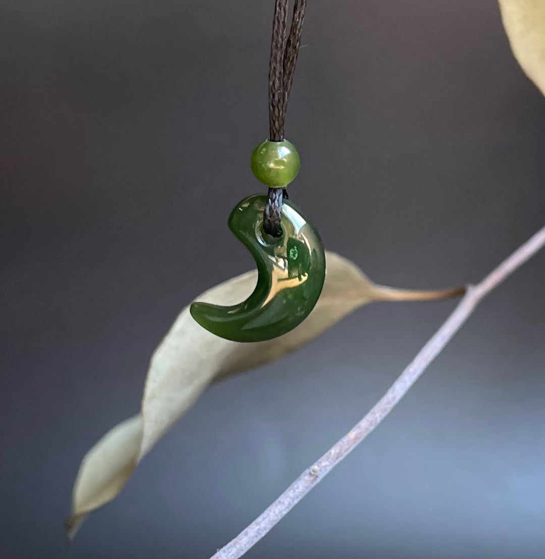 Jade magatama pendant on a dark cord with bead, hanging with a leaf and soft neutral background
