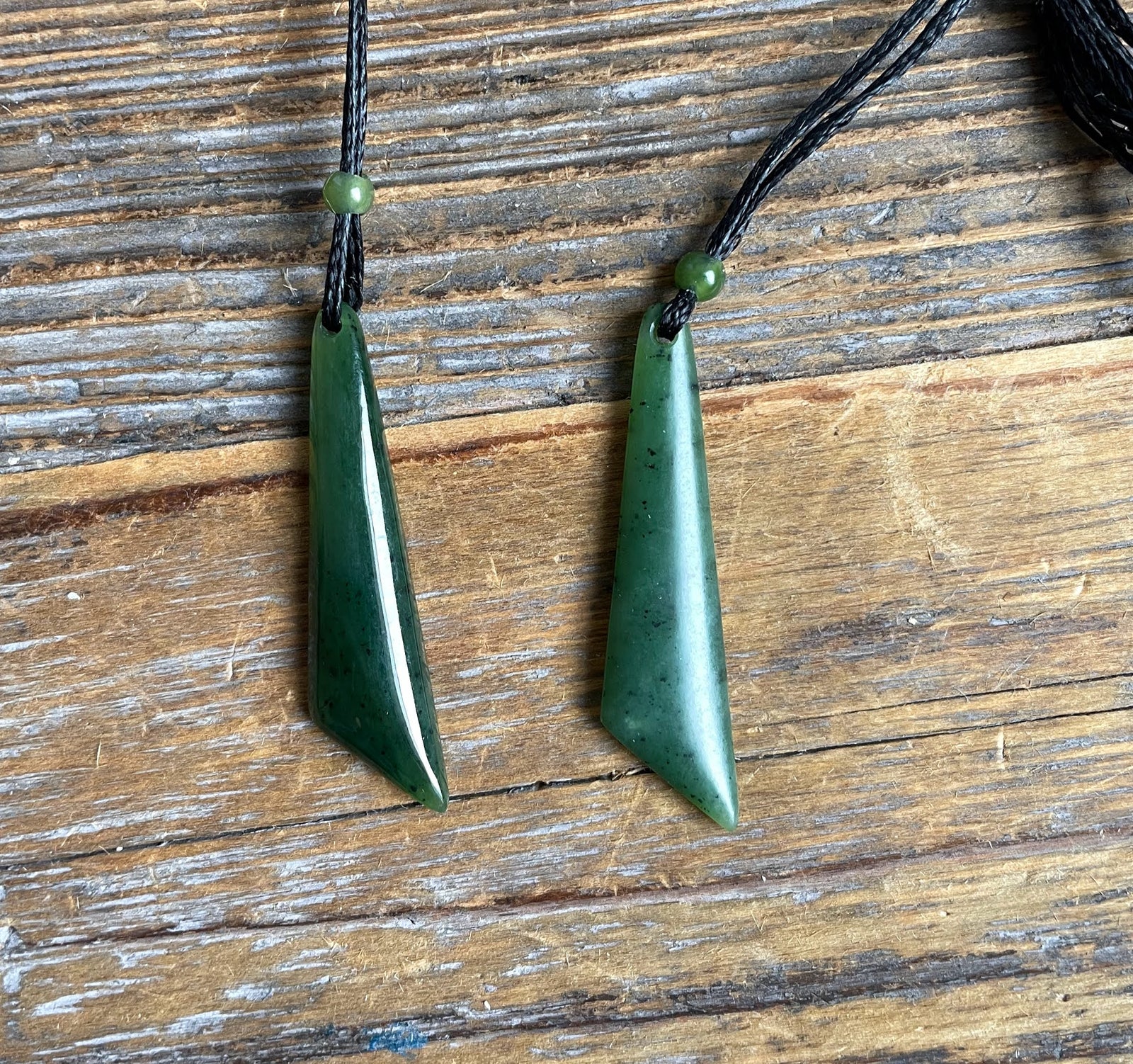 Two green jade drop pendants on black cords with small green beads against a wooden background