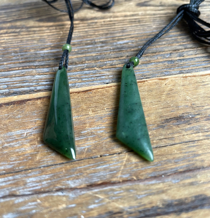 Two green jade drop pendants on black cords with beads placed on a wooden surface