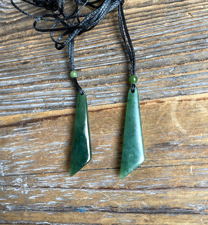 Two green jade drop pendants on braided black cords with green beads above on wood