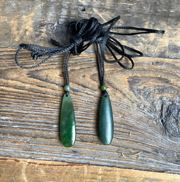 Two jade drop pendants with small beads on black cords laid on a rustic wooden surface.