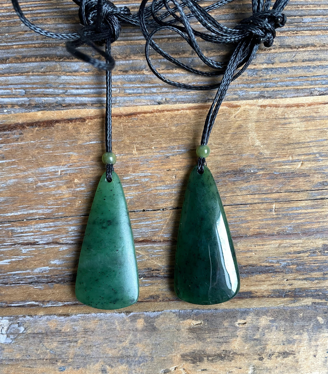 Two green jade teardrop pendants on black cords with small beads, arranged side by side on a wooden surface