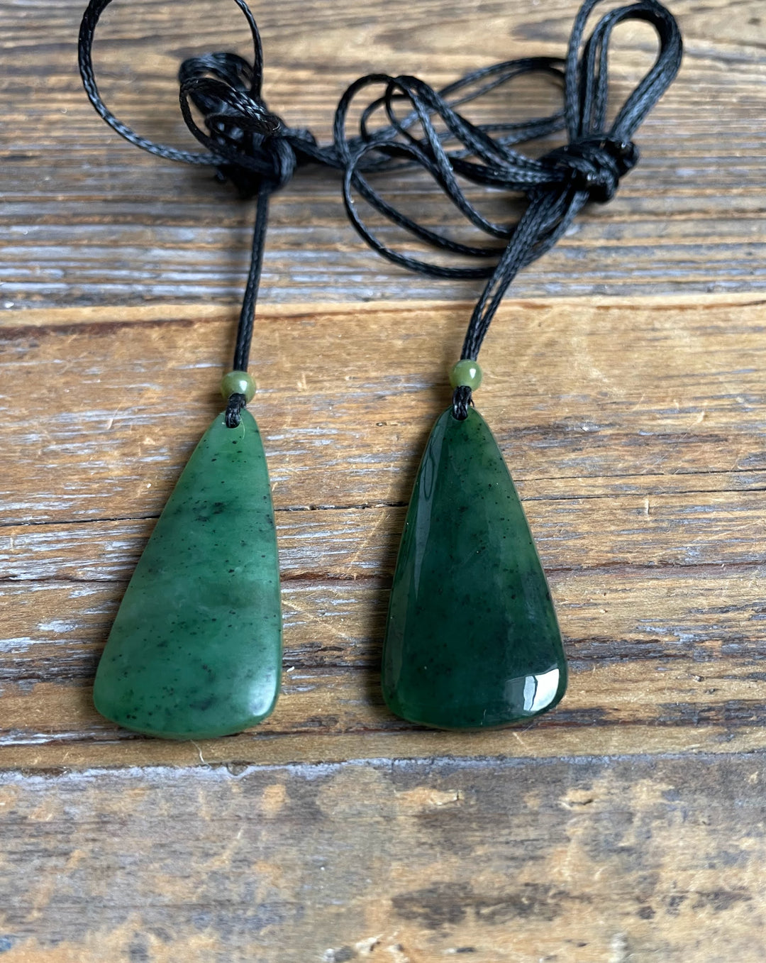 Pair of green jade drop pendants on black cords with small green beads against a wood background