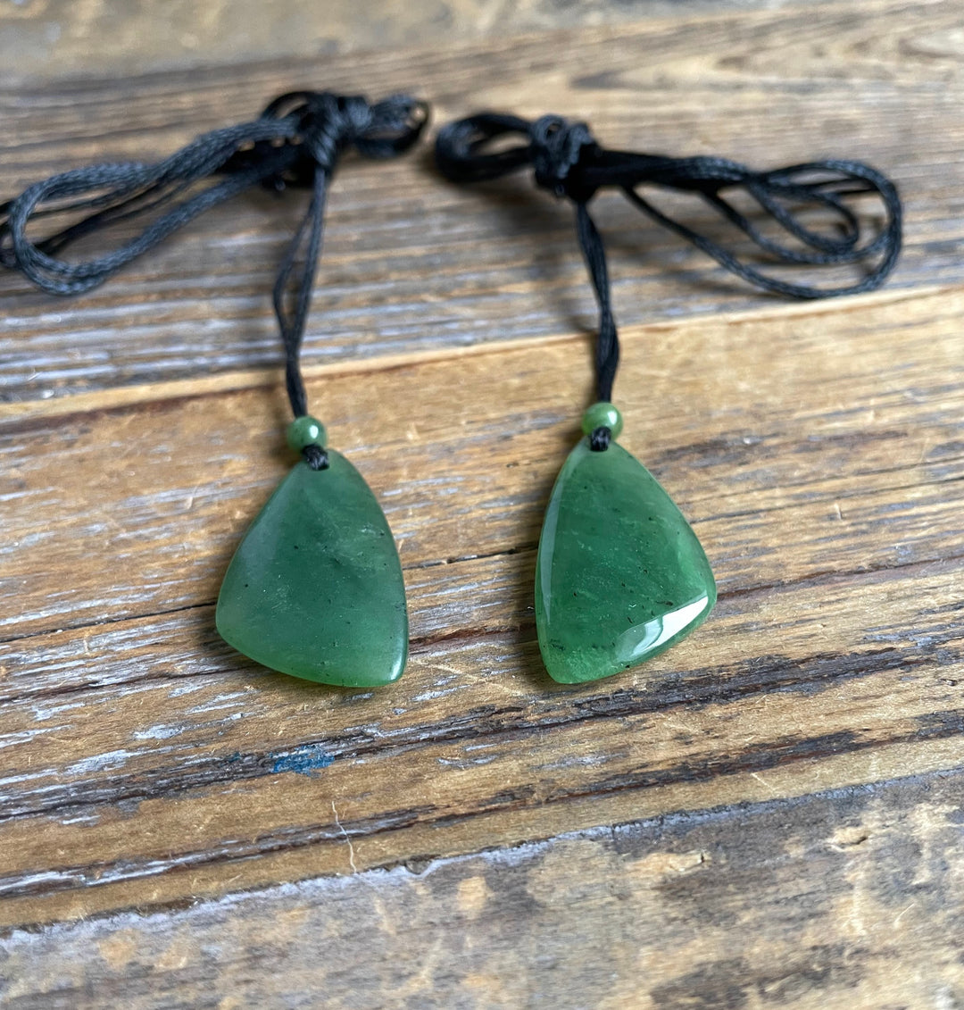 Two green jade teardrop pendants on black cords with matching beads resting on a wooden surface.