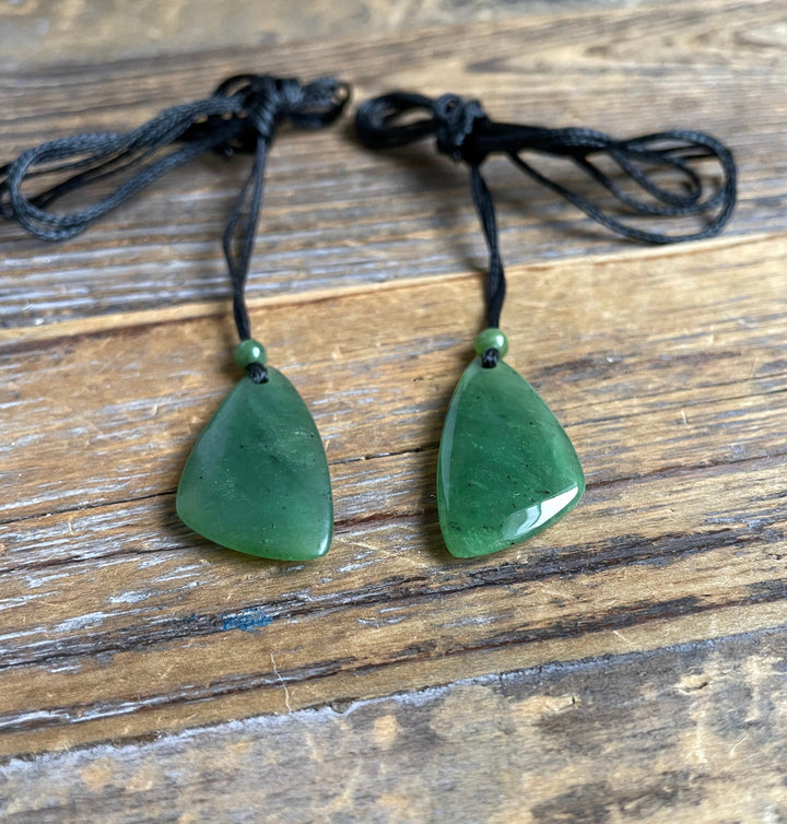 Two green jade teardrop pendants on black cords with matching beads resting on a wooden surface.