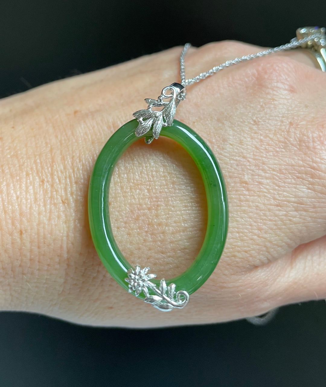 Jade green oval pendant on a silver chain worn on the wrist, featuring leaf and floral silver accents.