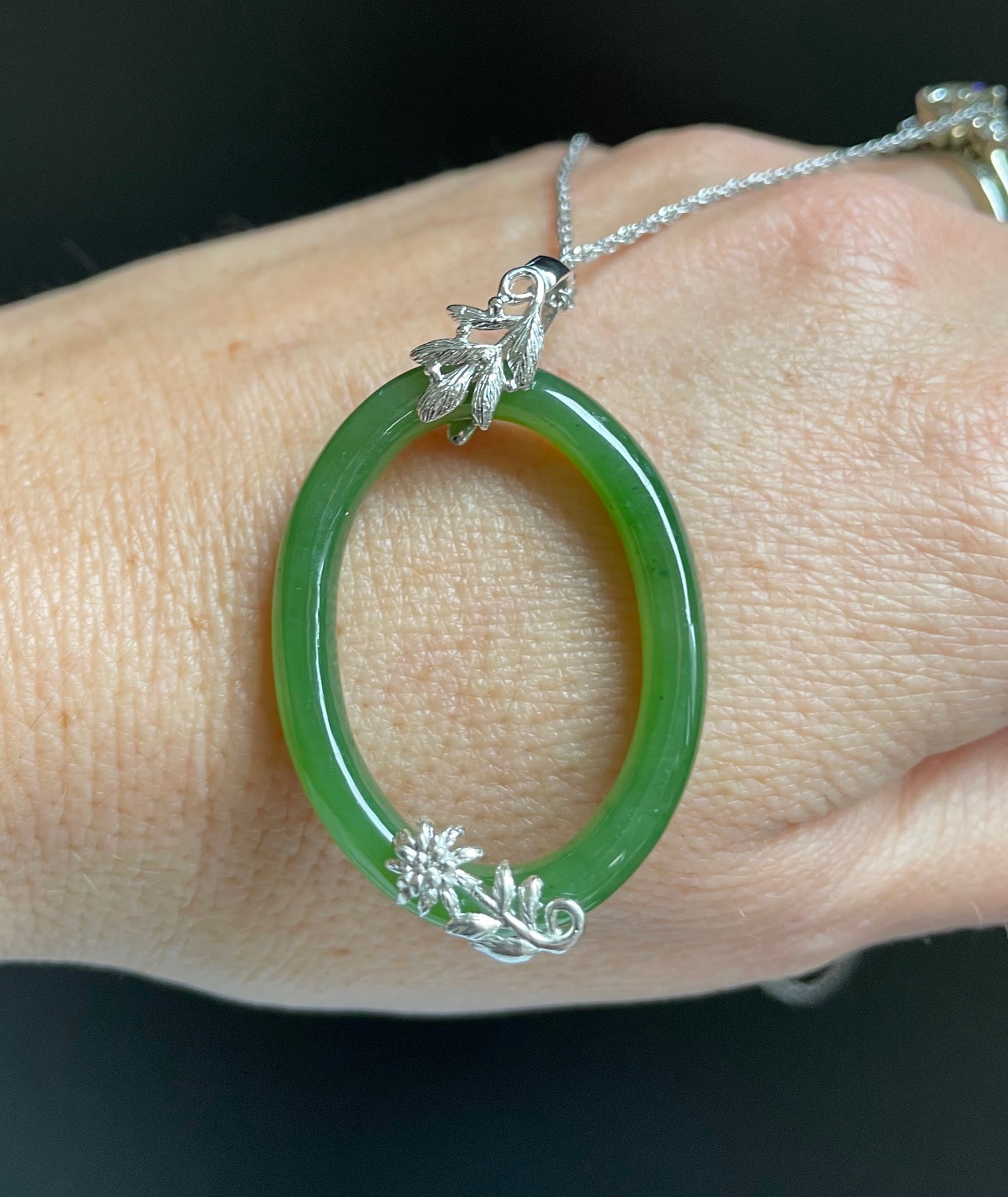 Jade green oval pendant on a silver chain worn on the wrist, featuring leaf and floral silver accents.