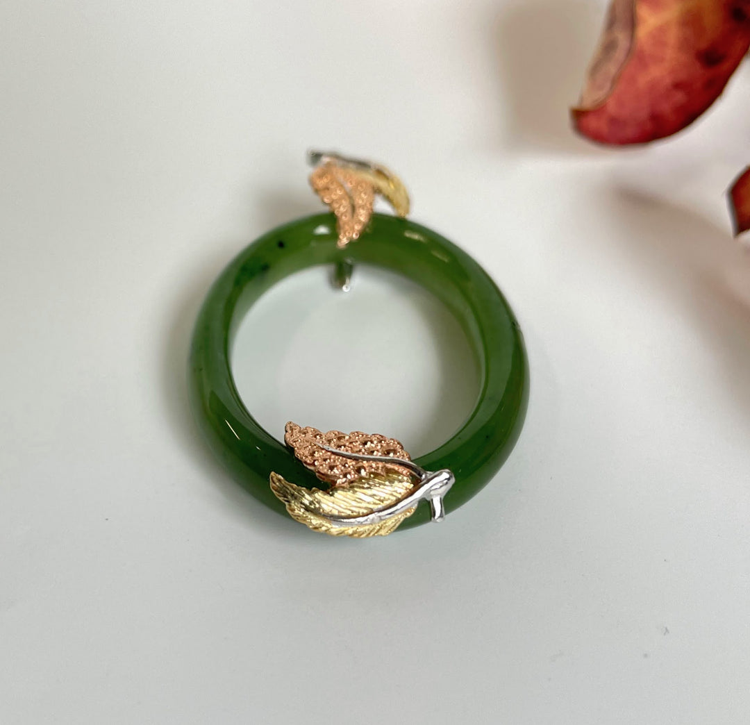 Green jade oval pendant with rose gold leaf accents on the bottom, displayed on a white surface.