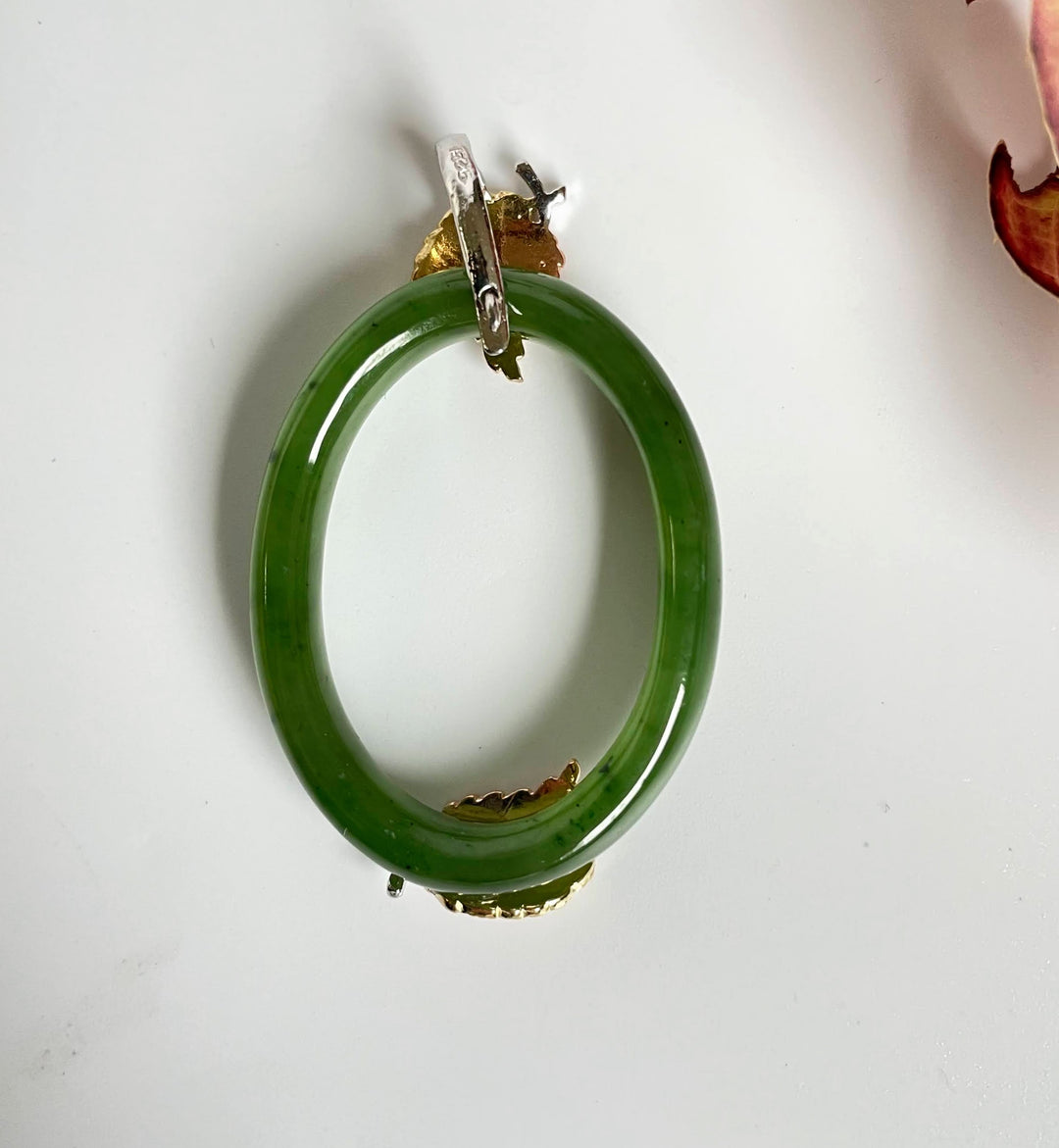 Green jade oval pendant with gold leaf accents and a silver bail, shown on a white background.