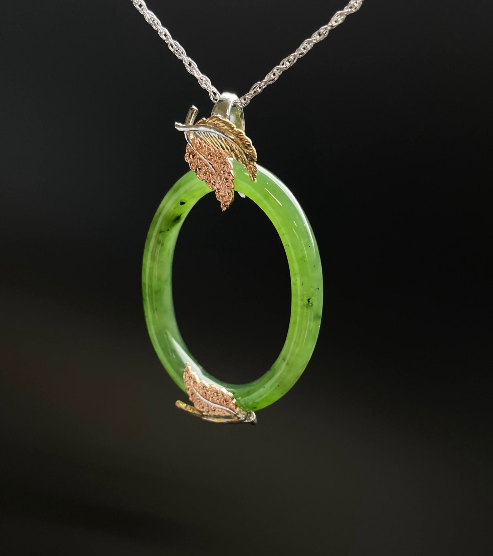 Green jade oval pendant with gold and copper leaf accents on a silver chain.