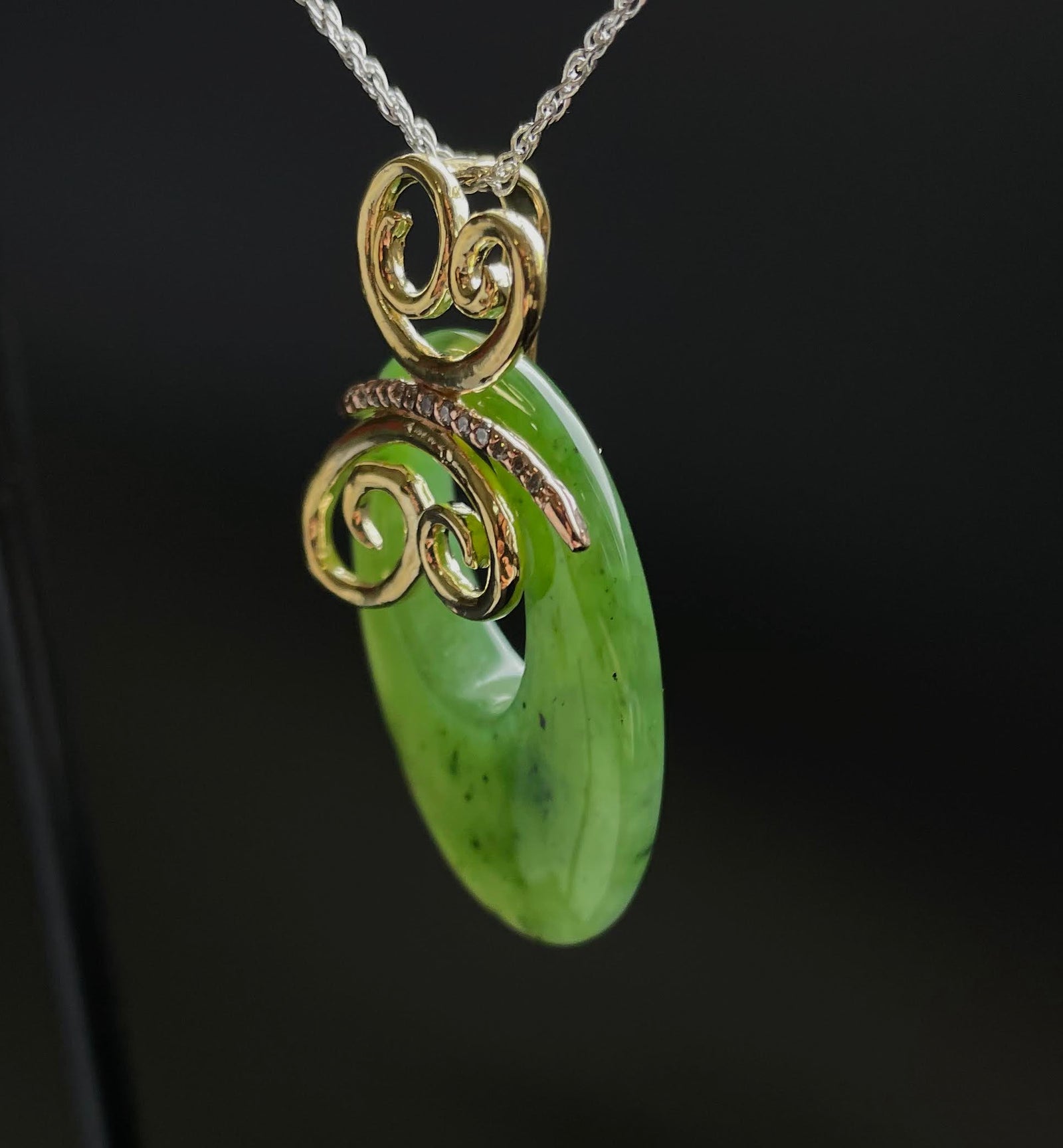 Close up of green jade oval pendant with gold scrollwork on a silver chain against a dark background.