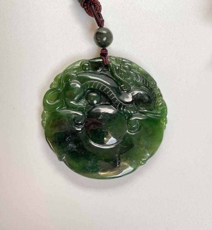 Double sided dragon pendant 50mm carved from green jade circular disc on a burgundy cord.