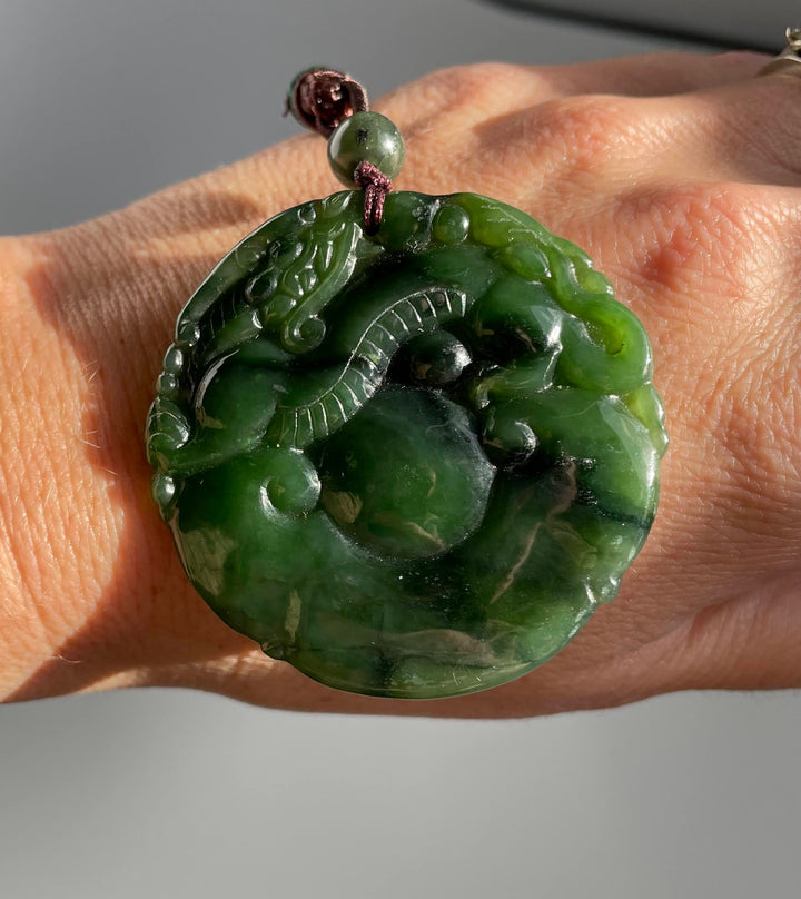 Green jade circular dragon pendant held on a hand with a burgundy braided cord above.