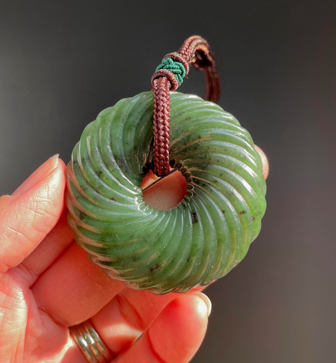 Close up of round green jade twist pendant hanging from a braided brown cord, held between fingers.