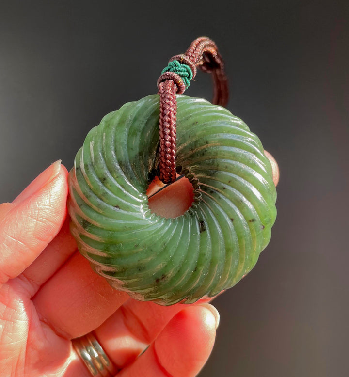 Close up of round green jade twist pendant hanging from a braided brown cord, held between fingers.