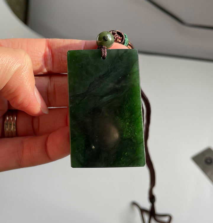 Dark green rectangular jade pendant with brown cord and green bead held in a hand.