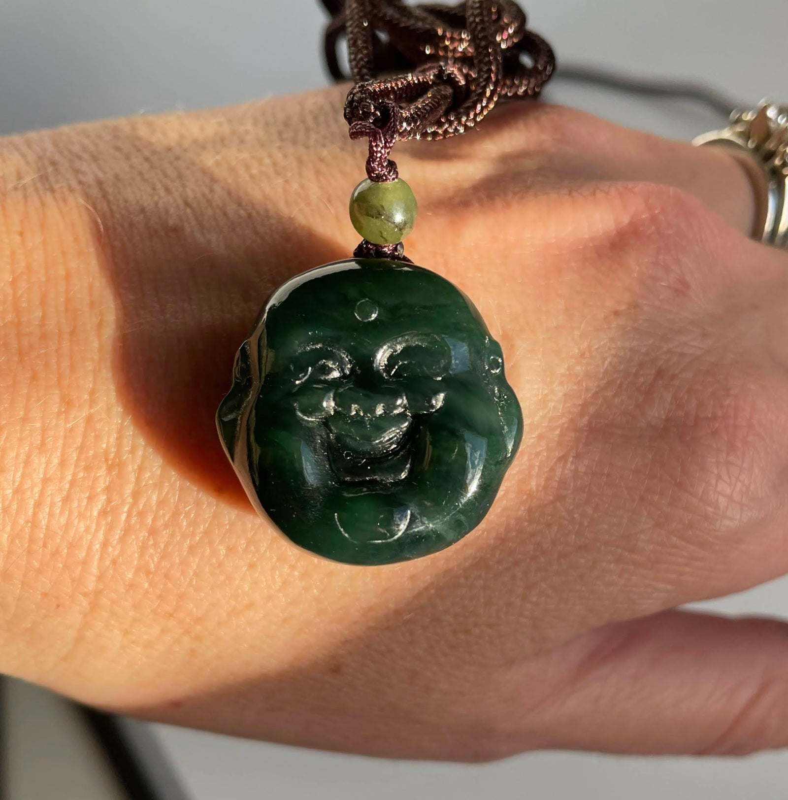 Close up of a Happy Buddha Head pendant carved from dark green stone on a brown braided cord with a small green bead