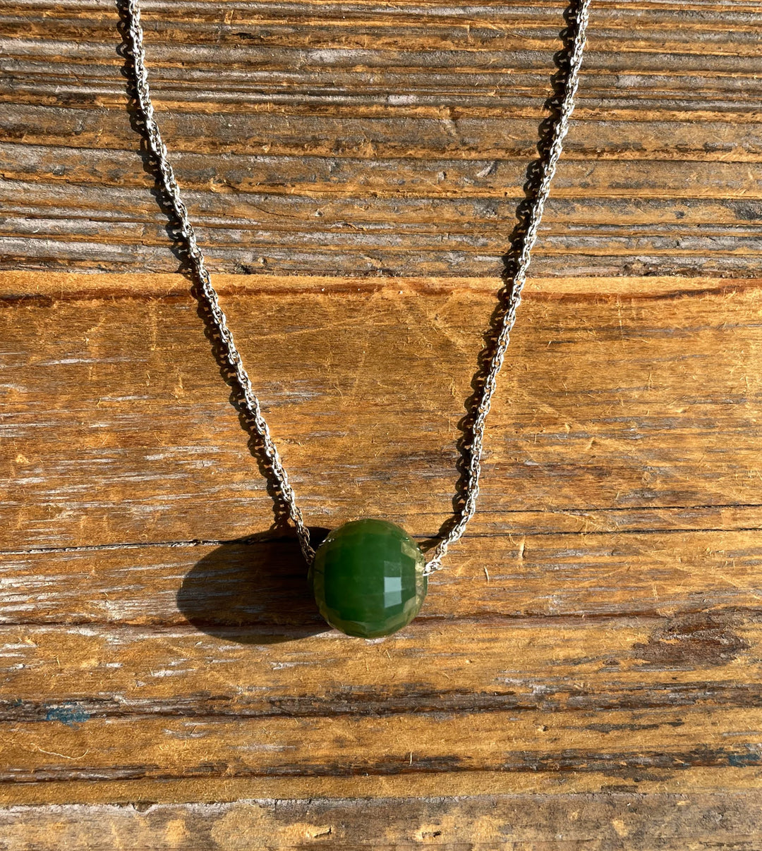 Green faceted jade bead pendant on a silver chain against a rustic wooden background.