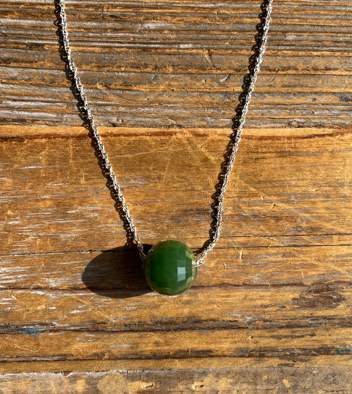 Green faceted jade bead pendant on a silver chain against a rustic wooden background.
