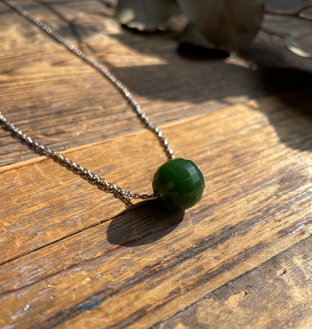 Close-up of a green faceted jade bead pendant on a silver chain resting on a wooden surface.
