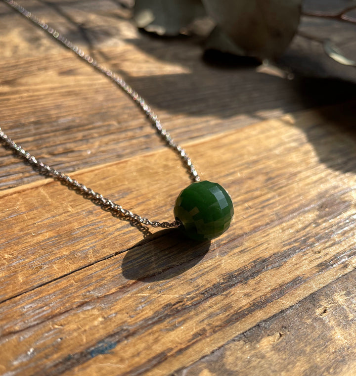 Close-up of a green faceted jade bead pendant on a silver chain resting on a wooden surface.