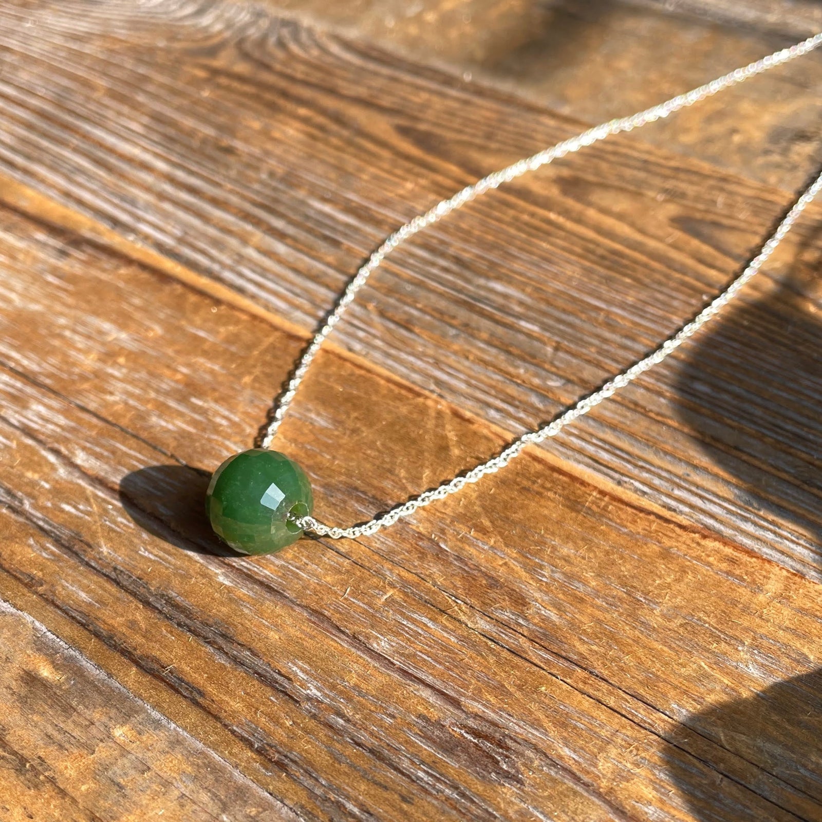 12mm faceted jade bead pendant on a silver chain resting on a wooden surface