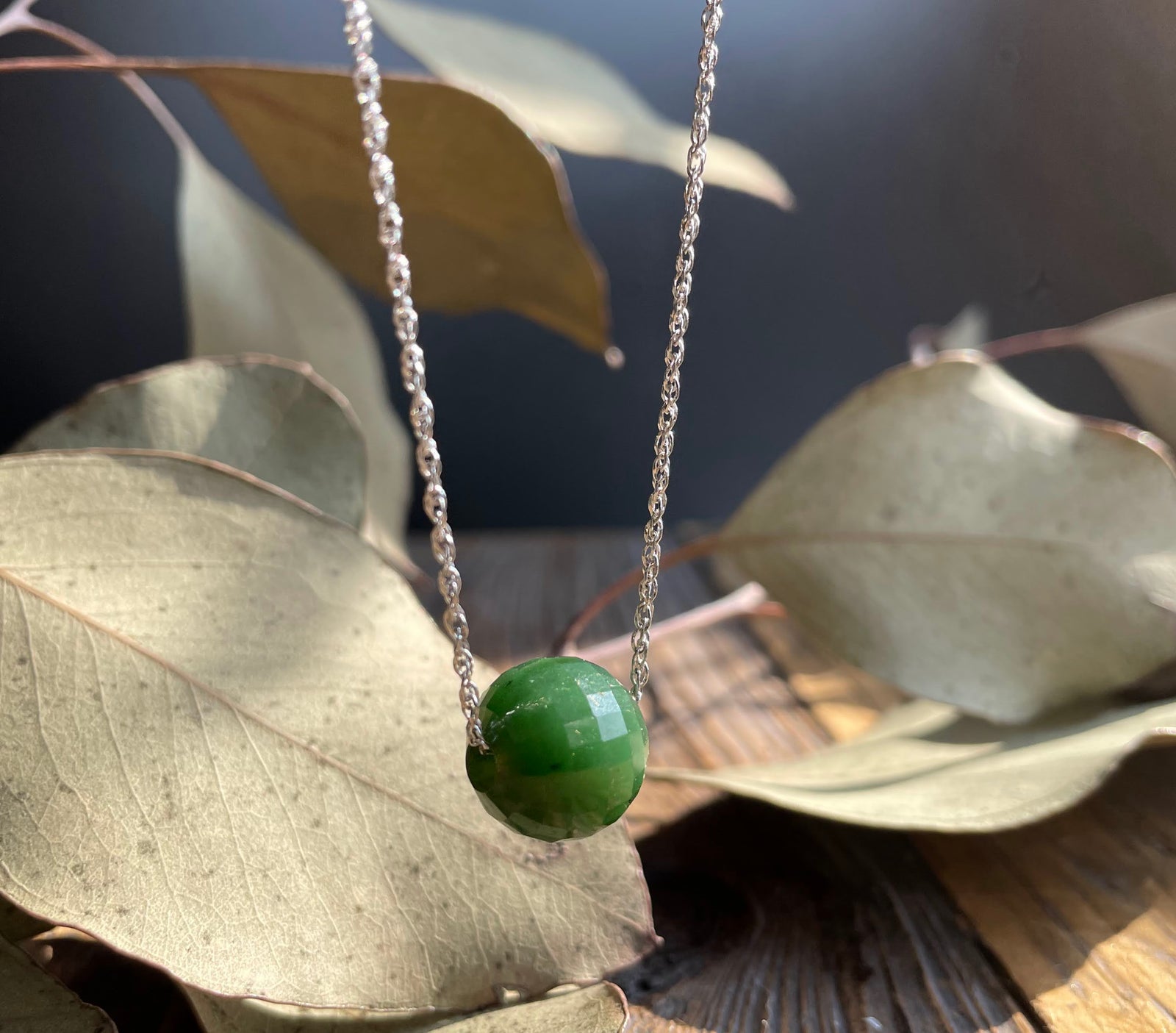 Green faceted jade bead pendant on a silver chain against dried leaves