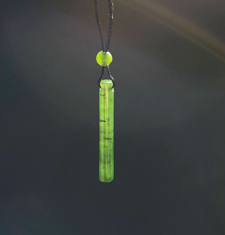 Jade cylinder pendant on a black cord with a round bead, shown against a dark blurred background